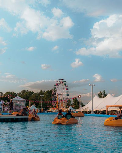 Festival Parasol - Des personnes font du pédalo dans une grande piscine extérieure, avec une grande roue et des tentes blanches en arrière plan