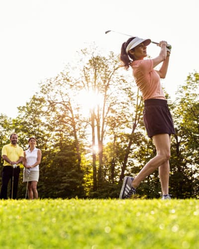 A woman plays golf, with three other individuals observing her in the background.