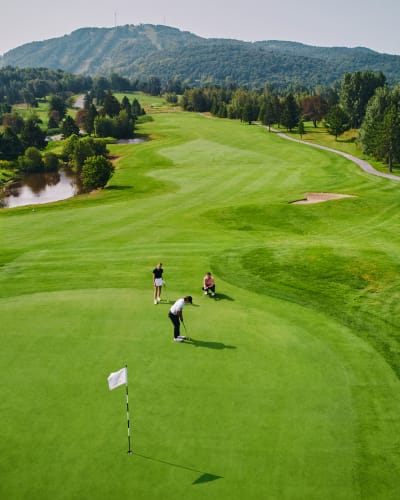 Trois personnes sur un vert au Golf Château-Bromont, avec la montagne en arrière-plan.