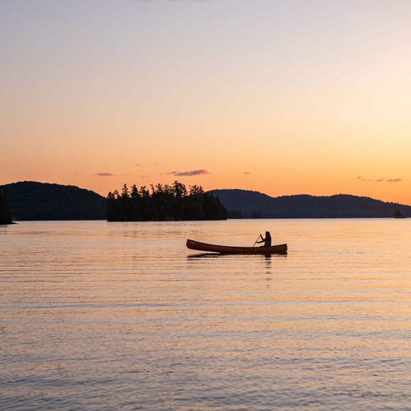Une personne est assise dans un canot sur l'eau, avec un coucher de soleil en arrière-plan