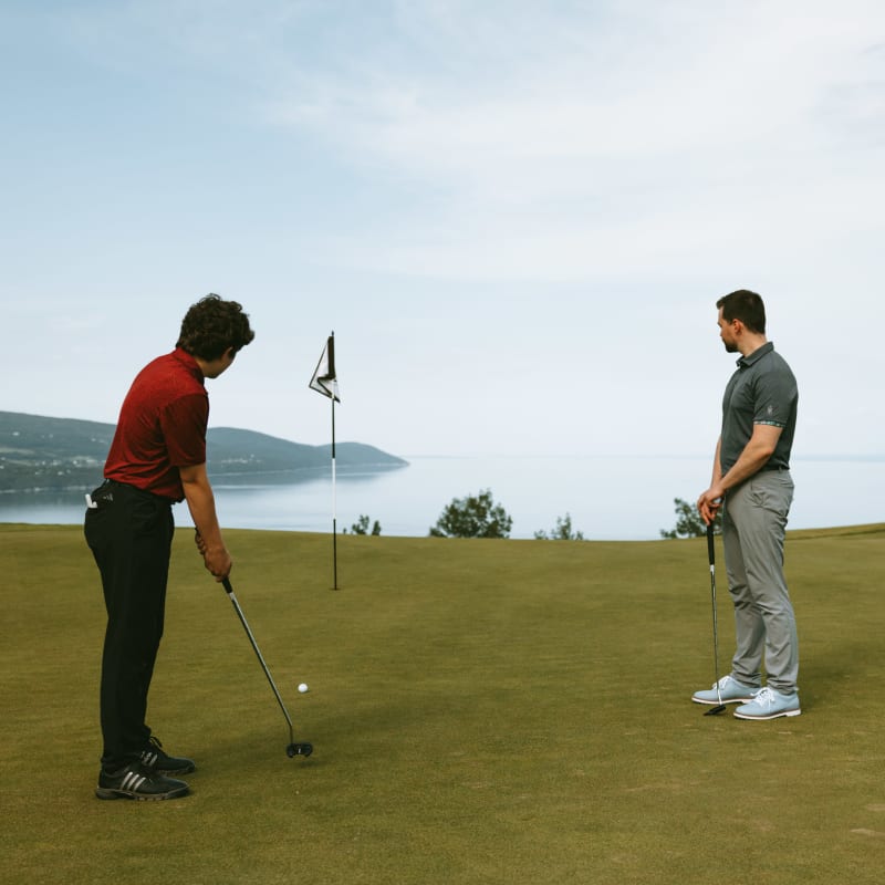 Jeune homme qui s'apprête à jouer un coup au Golf Manoir Richelieu, son père le regarde et le fleuve s'étend derrière eux.