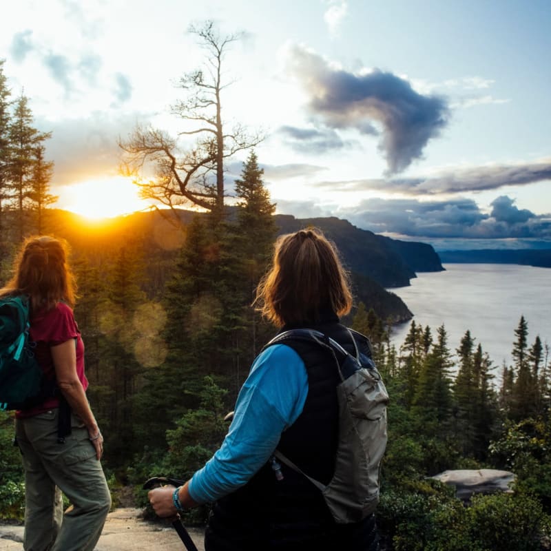 Parc national du Fjord-du-Saguenay - Deux femmes en haut d'une montagne, la forêt et le couché de soleil en arrière-plan.