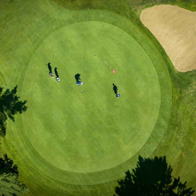 Vue aérienne d'un vert de golf bordé d'une fosse de sable; on y voit 4 golfeurs et leurs ombres.