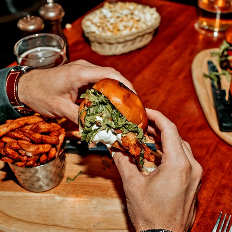 Deux mains tenant un hamburger au-dessus d'une planche avec des frites sur une table de restaurant.