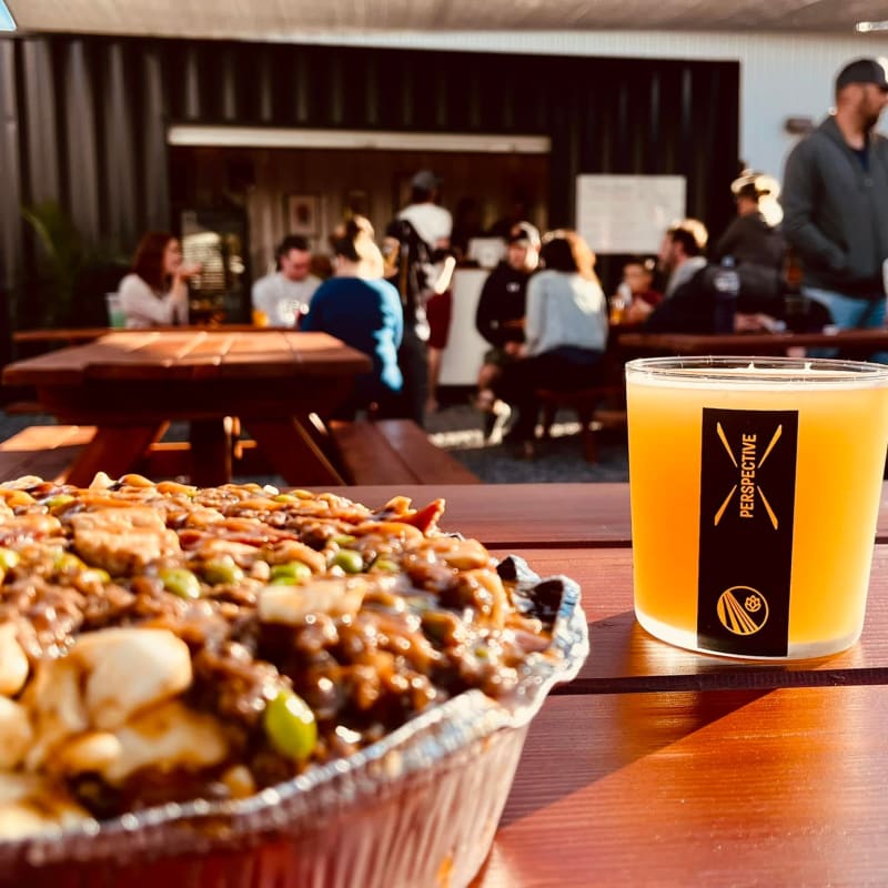 Gros plan sur une poutine et une bière; à l'arrière, des gens profitent de la terrasse de la microbrasserie Perspective.